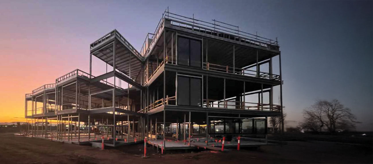Low-angle view of a multi-story modular prefabricated steel building frame under construction at dusk, featuring open steel framing, glass windows, and safety scaffolding against a purple and orange gradient sky.