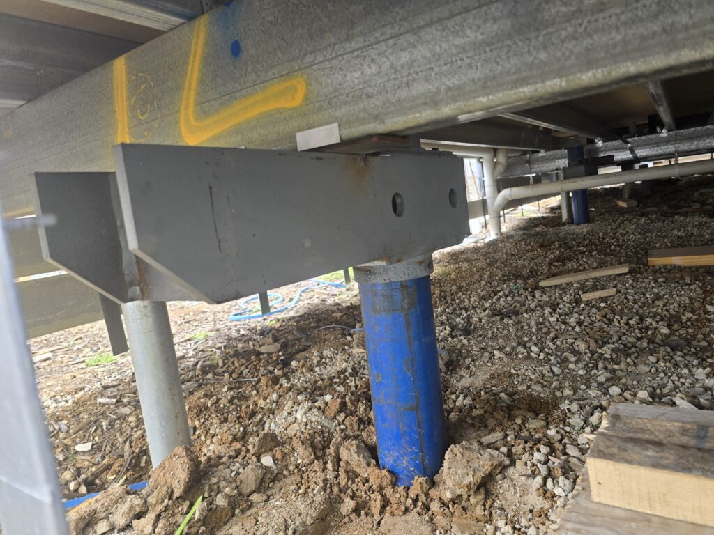 Low-angle view under a building structure showing a blue steel screw pile attached to a heavy galvanized support bracket, supporting the structural steel floor joists above an unpaved crawl space.