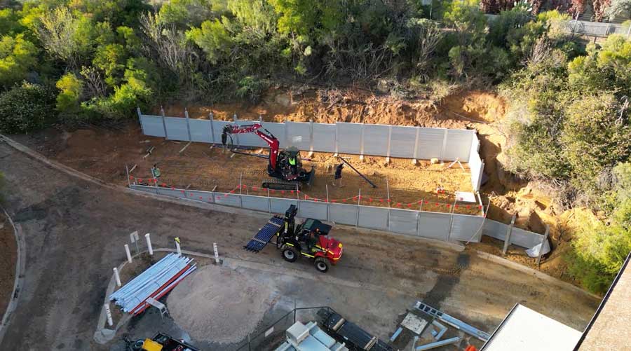 High aerial view of a civil works project showing a small excavator drilling screw piers inside a cleared earth plot enclosed by modern modular acoustic or retaining wall panels, with a all-terrain forklift nearby and raw construction materials stacked on a dirt roadway.