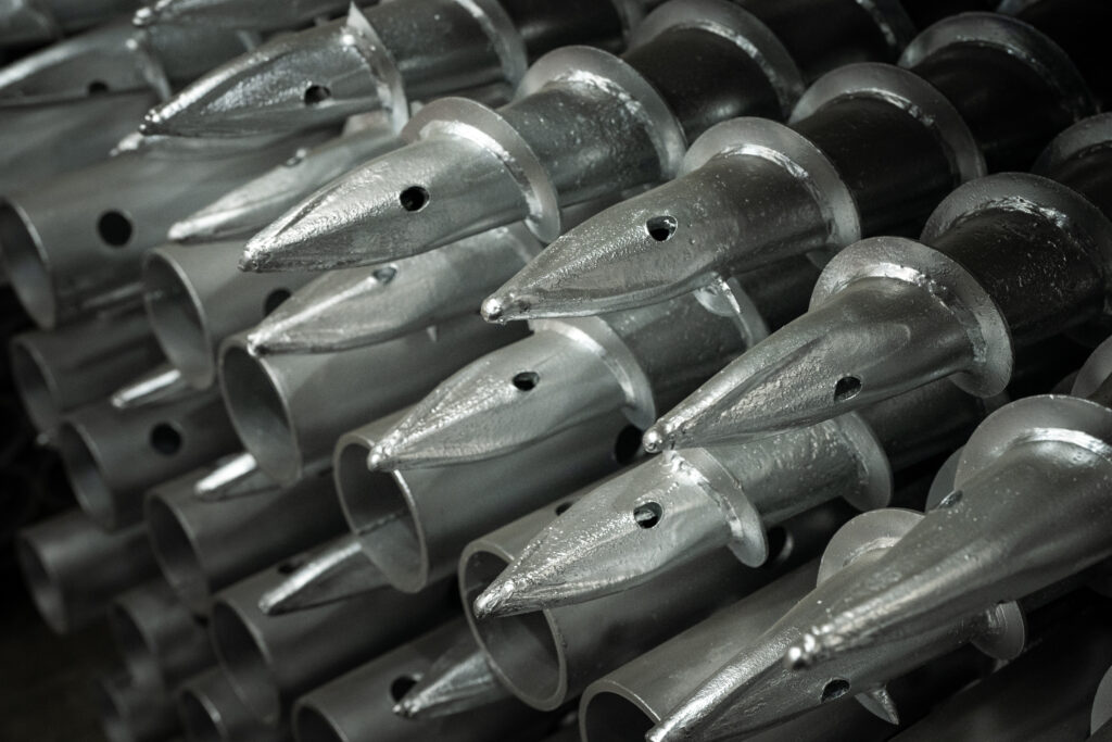 Macro view of a bundle of heavy-duty galvanized steel ground screws, highlighting the pointed tips, spiral helical threads, and pre-drilled pin holes on the pipe ends.