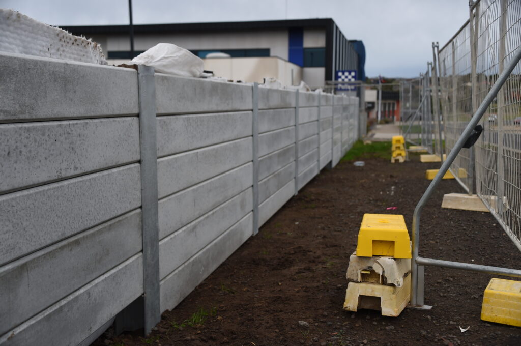 Ground-level view showing a long precast concrete sleeper retaining wall supported by steel H-beam posts, running parallel to temporary chain-link construction fencing with yellow counterweight feet.