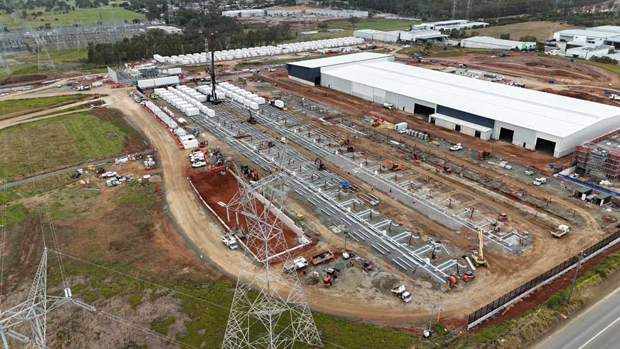 High-angle aerial view of an industrial battery energy storage site under construction, showing long rows of white BESS container units, concrete foundations, power transmission towers, and heavy construction equipment alongside a large industrial warehouse.