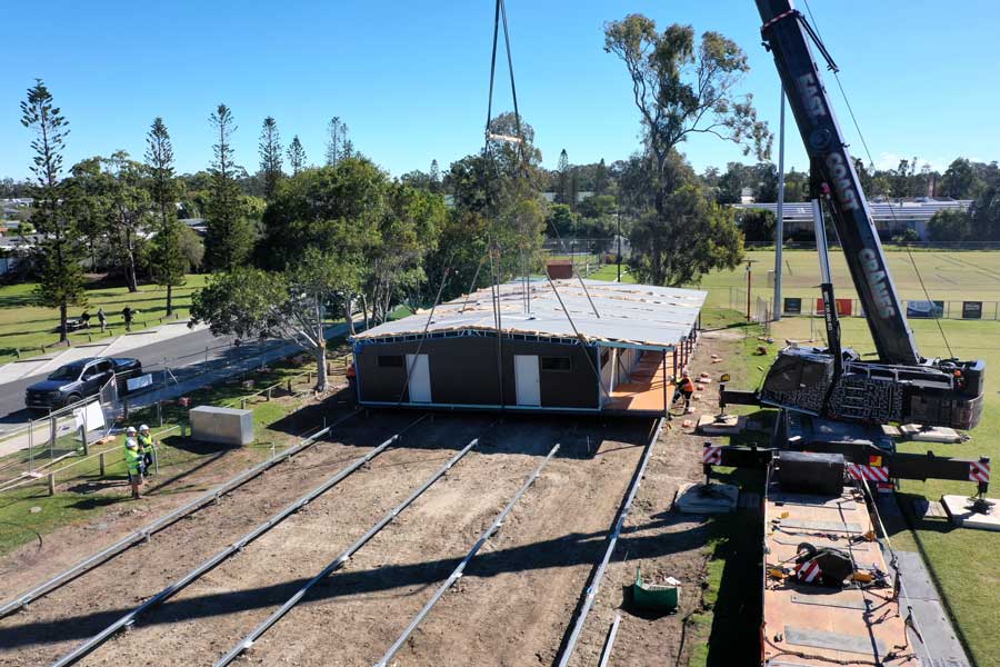 High-angle outdoor view of an East Coast Cranes mobile crane lifting and lowering a long, single-story prefabricated modular building section onto parallel steel foundation rails on a clear, sunny day.