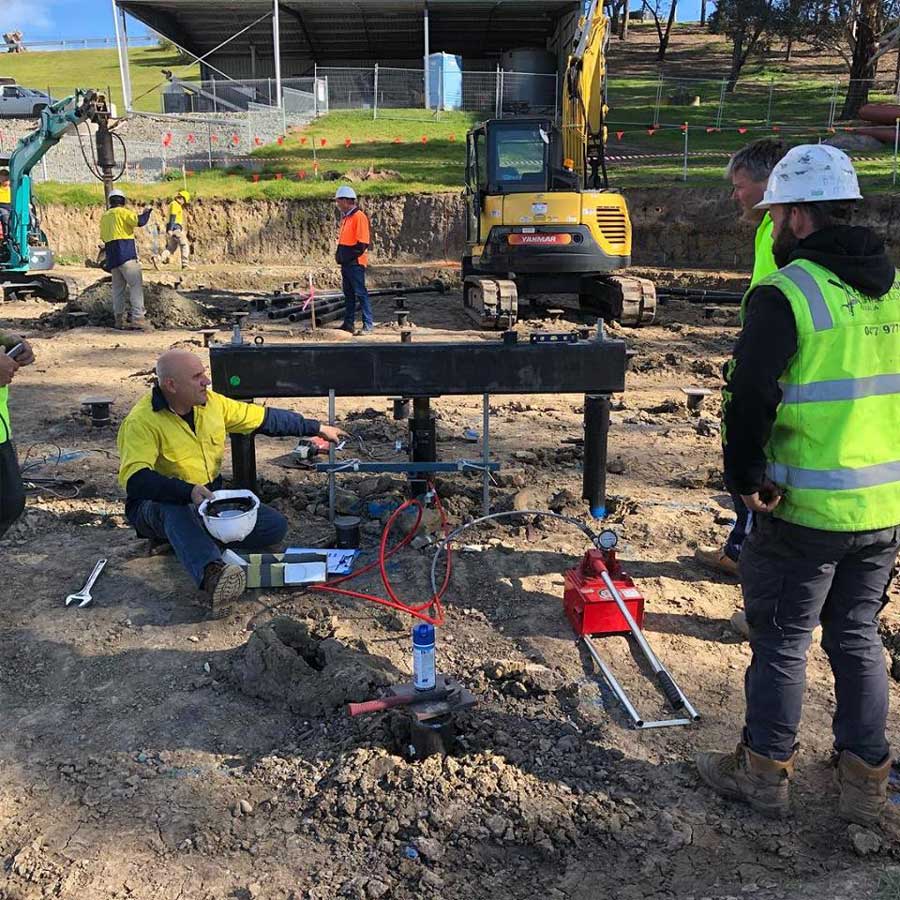 Outdoor view of engineers and construction workers performing a static load test on foundation screw piles, featuring a heavy steel reaction beam, hydraulic jacks, gauges, and excavators in the background.