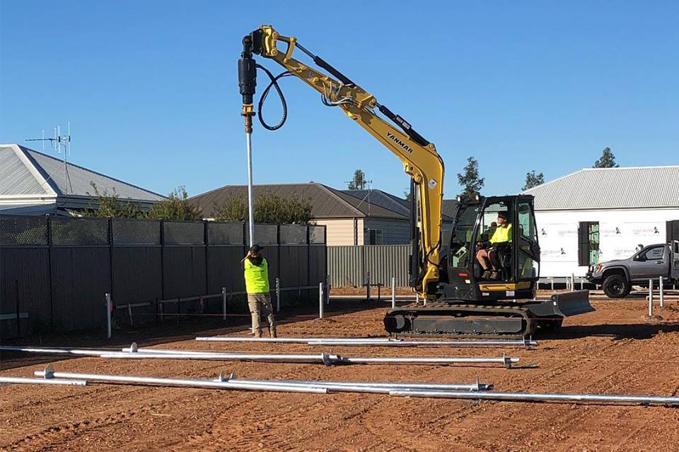 Outdoor view of a yellow mini excavator with a hydraulic drive attachment drilling vertical steel screw piles into red dirt, guided by a worker in high-visibility safety gear near suburban homes.