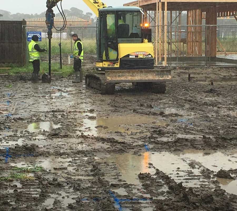 Outdoor view of a yellow mini excavator operating in thick, wet mud, using a hydraulic drive to install vertical foundation screw piers with two construction workers in high-vis vests supervising nearby.