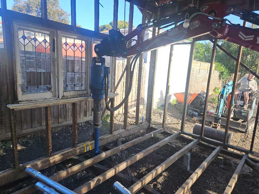 Interior construction view showing a machinery arm with a hydraulic drive driving a steel foundation screw pier into the ground through open floor joists, inside a partially renovated wooden house structure featuring leadlight windows.