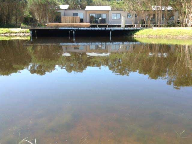 Wide view across calm water showing a wooden boardwalk deck anchored by steel screw pile foundations, positioned in front of a modern modular waterfront home surrounded by trees.