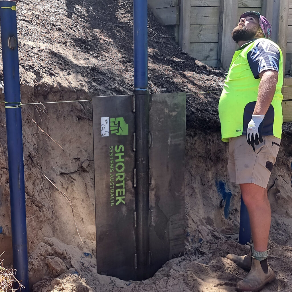 Construction worker in a high-visibility shirt standing near an excavated earth embankment, looking up as a steel Shortek Systems Australia shoring panel is driven into the ground along blue guide posts.