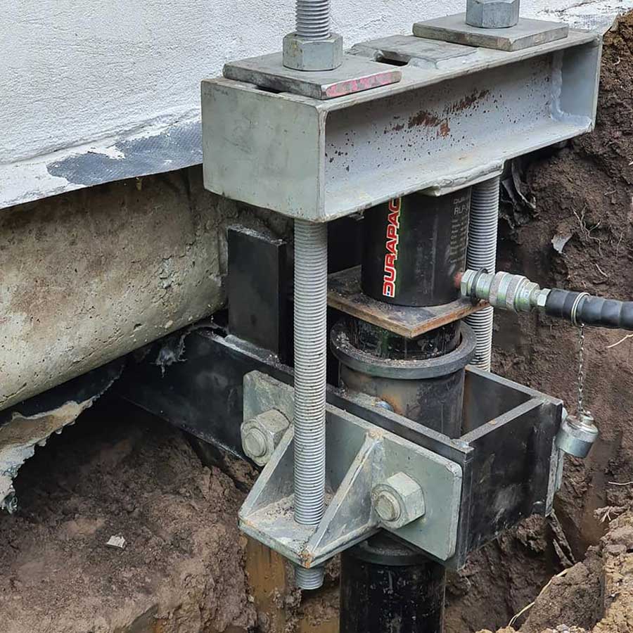 Extreme close-up of a Durapac hydraulic cylinder positioned inside a heavy-duty steel underpinning bracket assembly with threaded rods, lifting a concrete slab foundation from an excavated pit.