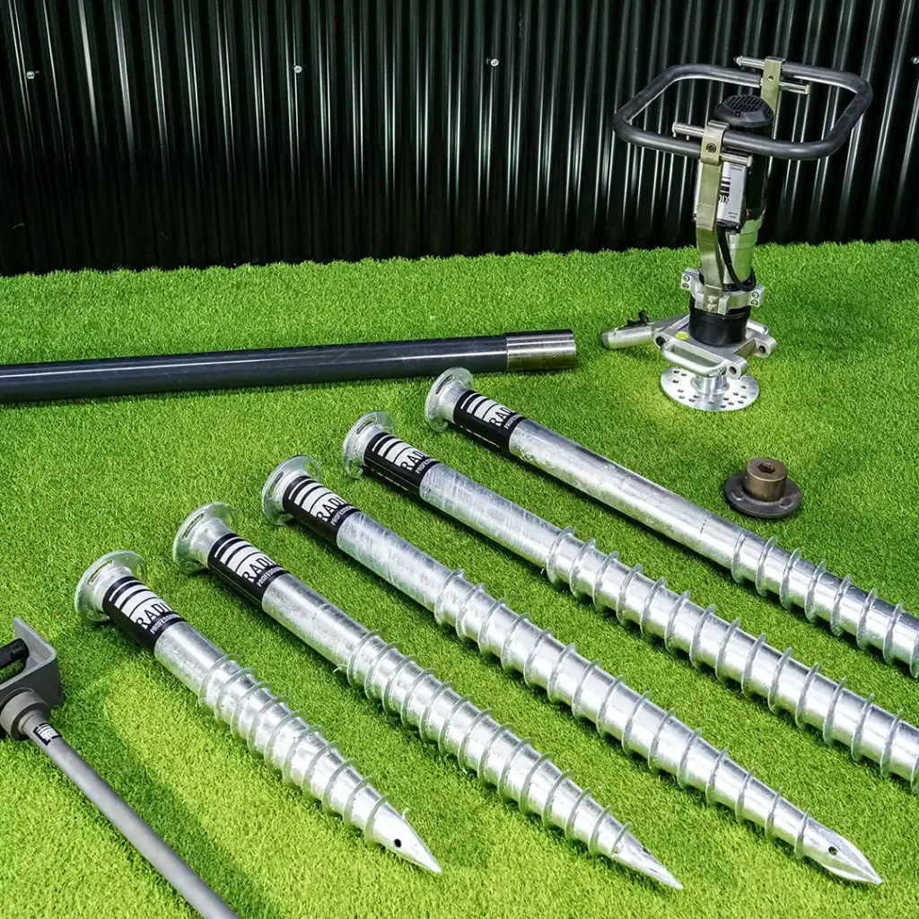 Overhead view of a RADIX ground screw installation kit displayed on artificial grass, including five galvanized steel ground screws of varying lengths, a handheld electric installation tool with a double-handled grip, and extension bars.