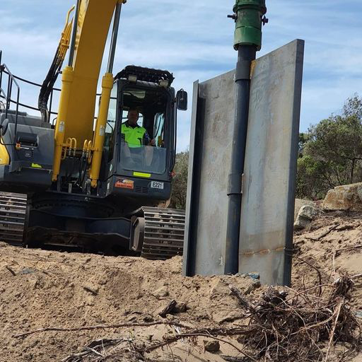 Yellow tracked excavator using a hydraulic drive attachment to press a heavy steel Shortek shoring panel vertically into a sandy dirt embankment on an outdoor construction site.